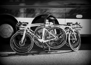 Monochromatická fotografie závodních bicyklů zaparkovaných před autobusem v kontextu sportovní akce.